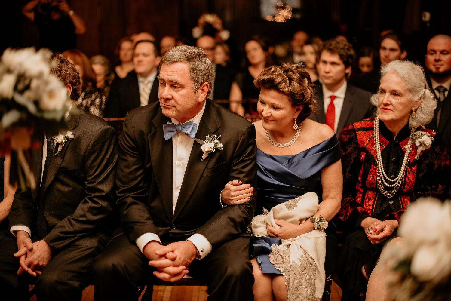 Wedding guests seated during ceremony, man in tuxedo with boutonniere beside elegantly dressed women.