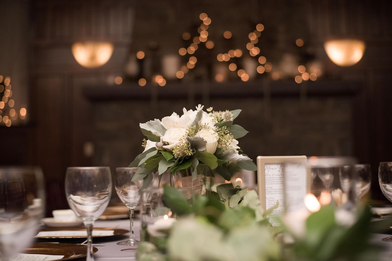 Elegant wedding reception table with white floral centerpiece, greenery, and glowing bokeh lights in a cozy venue.