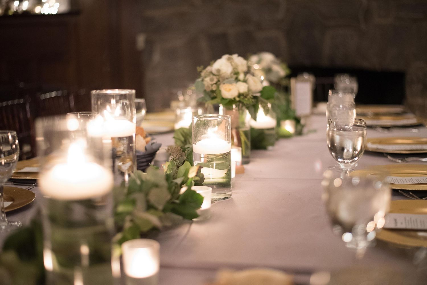 Elegant wedding reception table with white candles, greenery garland, and floral centerpieces in a warmly lit venue.
