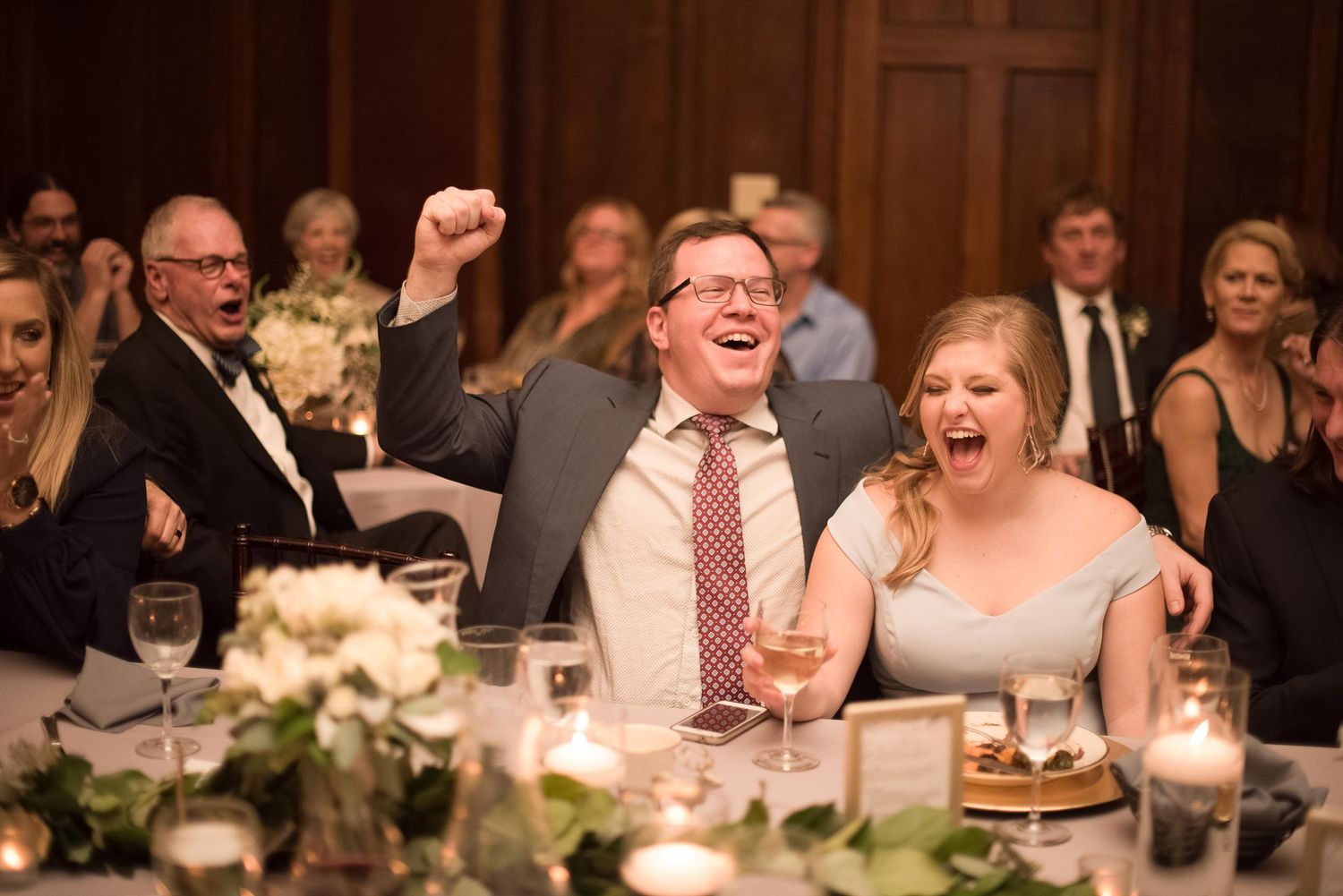 Guests laughing and cheering with fists raised at an elegant wedding reception dinner in a wood-paneled ballroom.