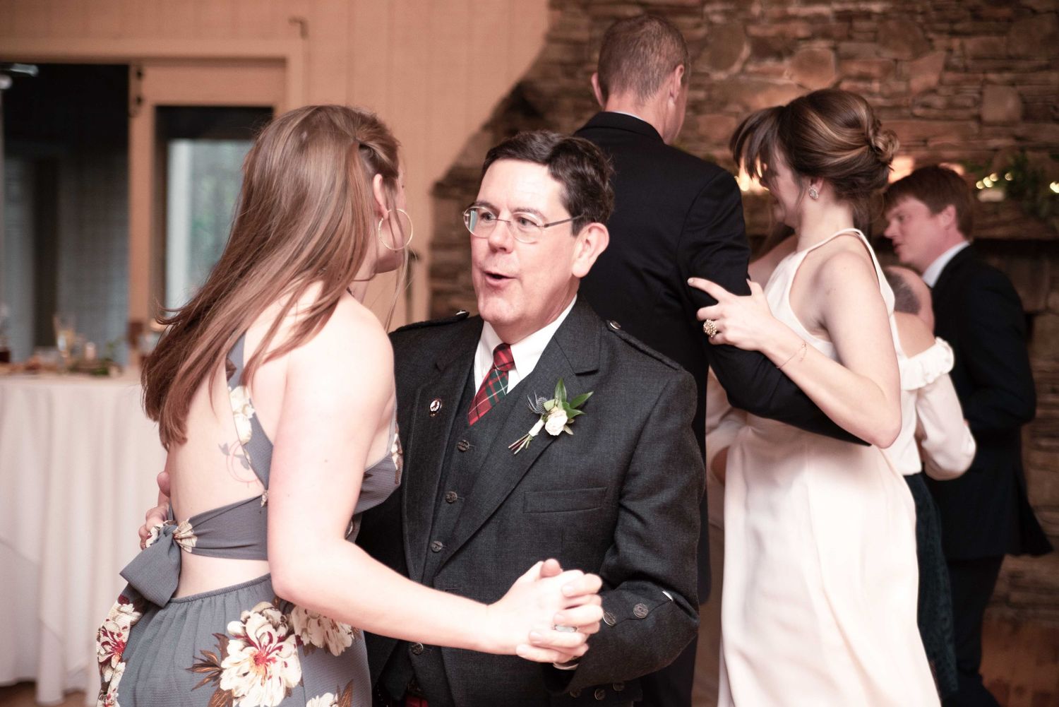 Guests dancing at a wedding reception in a rustic venue, man in dark suit with boutonniere dancing with woman.