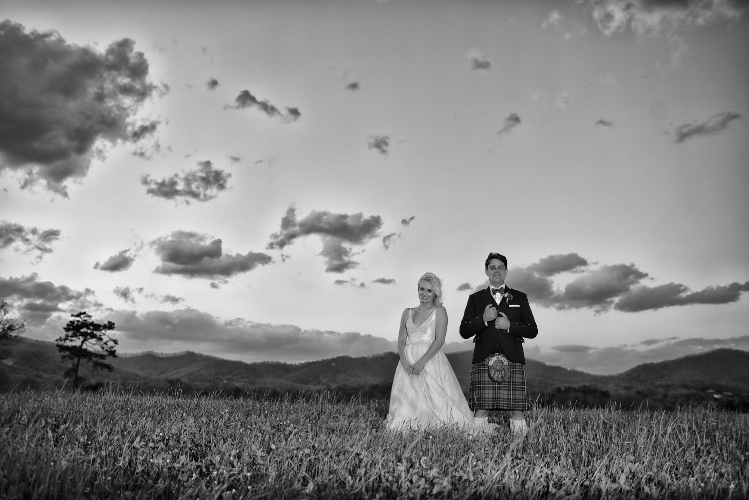 Black and white photo of a bride and groom standing in a field at sunset with mountains in the background.