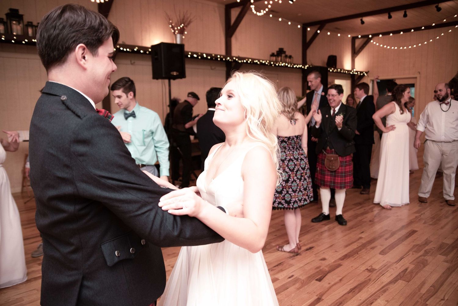 Bride and groom share their first dance at a rustic wedding reception with guests dancing in the background.