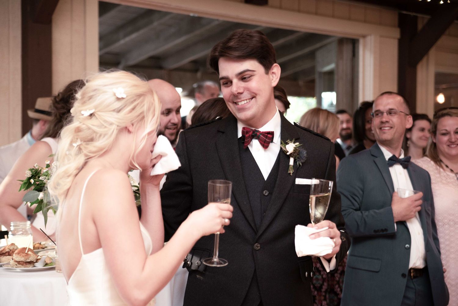 Groom in black suit with burgundy bow tie shares a toast with blonde bride during elegant wedding reception.