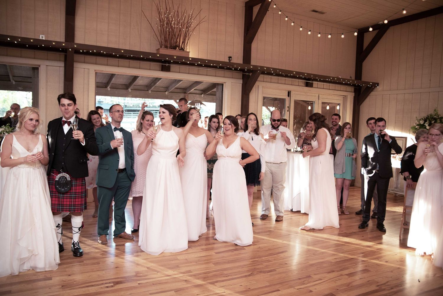 Wedding party in pink bridesmaid dresses and kilts raising glasses for a toast on a wooden dance floor.