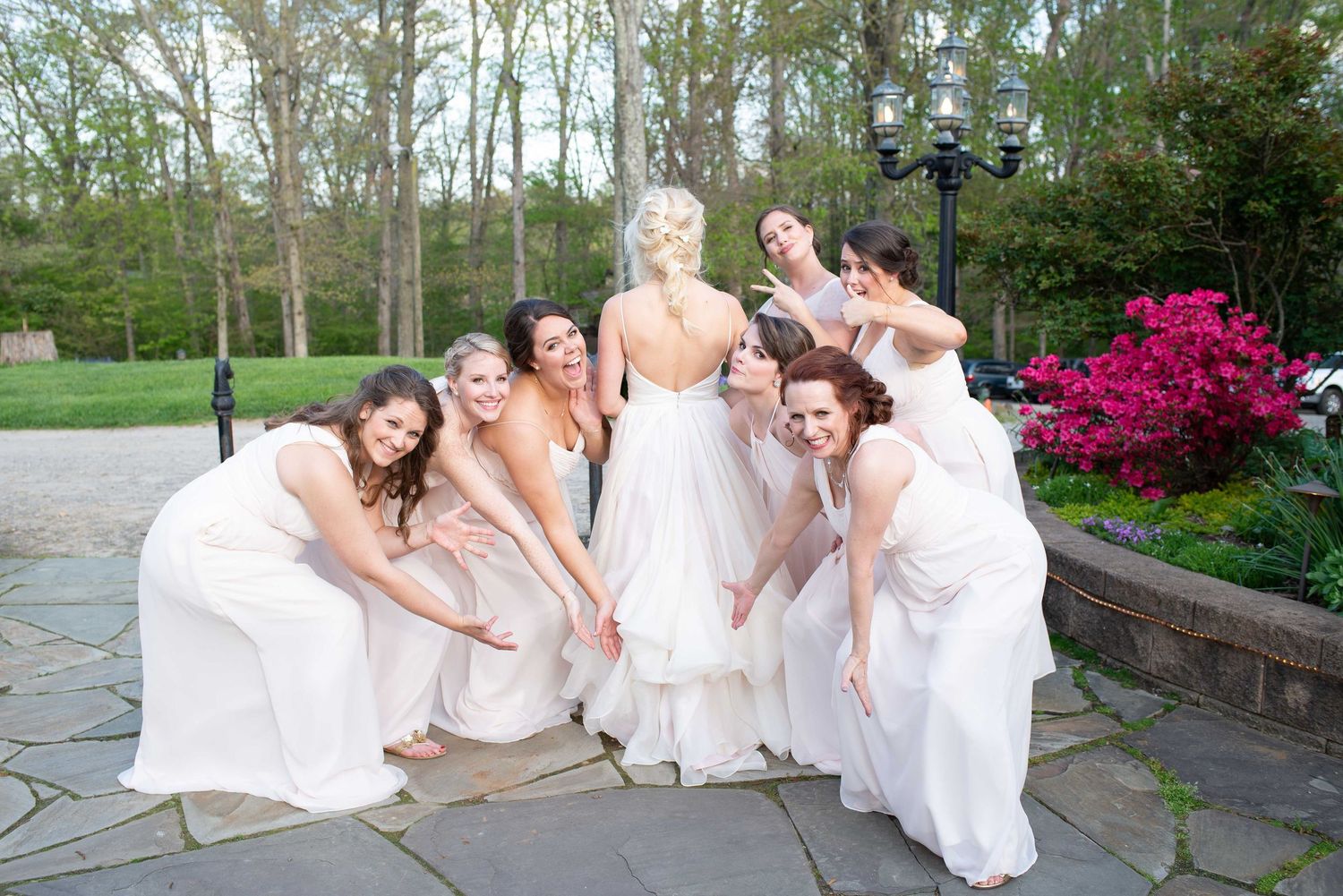 Bride in white gown surrounded by bridesmaids in white dresses posing on stone patio with blooming pink azaleas.