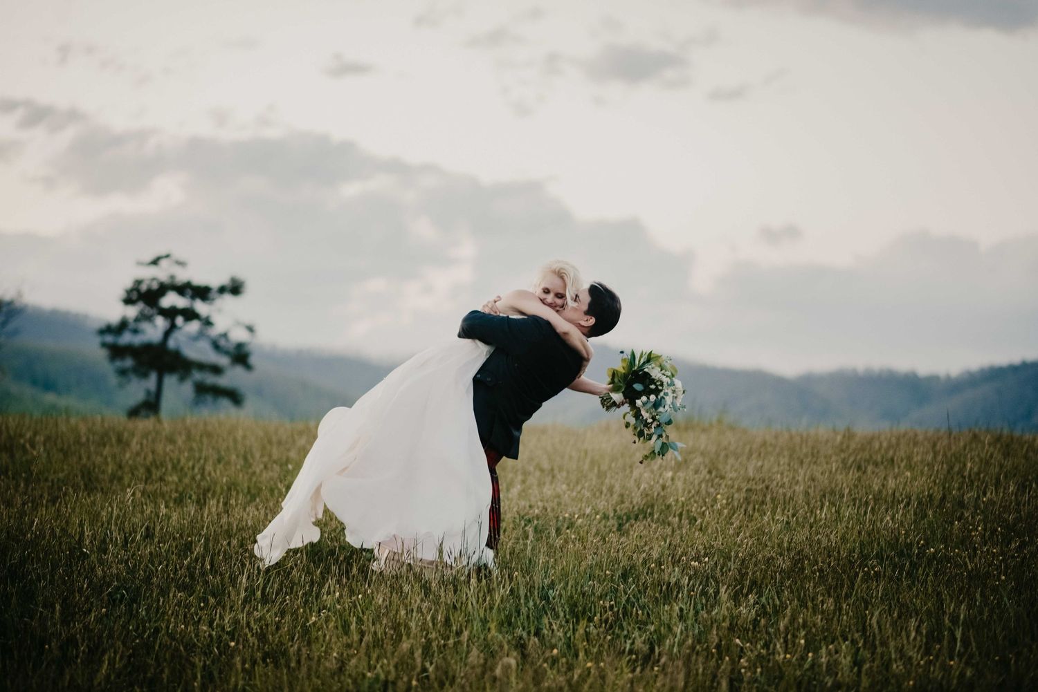 Groom dips bride for a romantic kiss in a lush green mountain meadow during outdoor wedding.