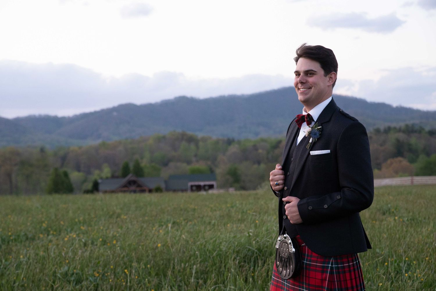 Smiling man in red tartan kilt and black jacket posing in scenic green field with misty mountains behind him.