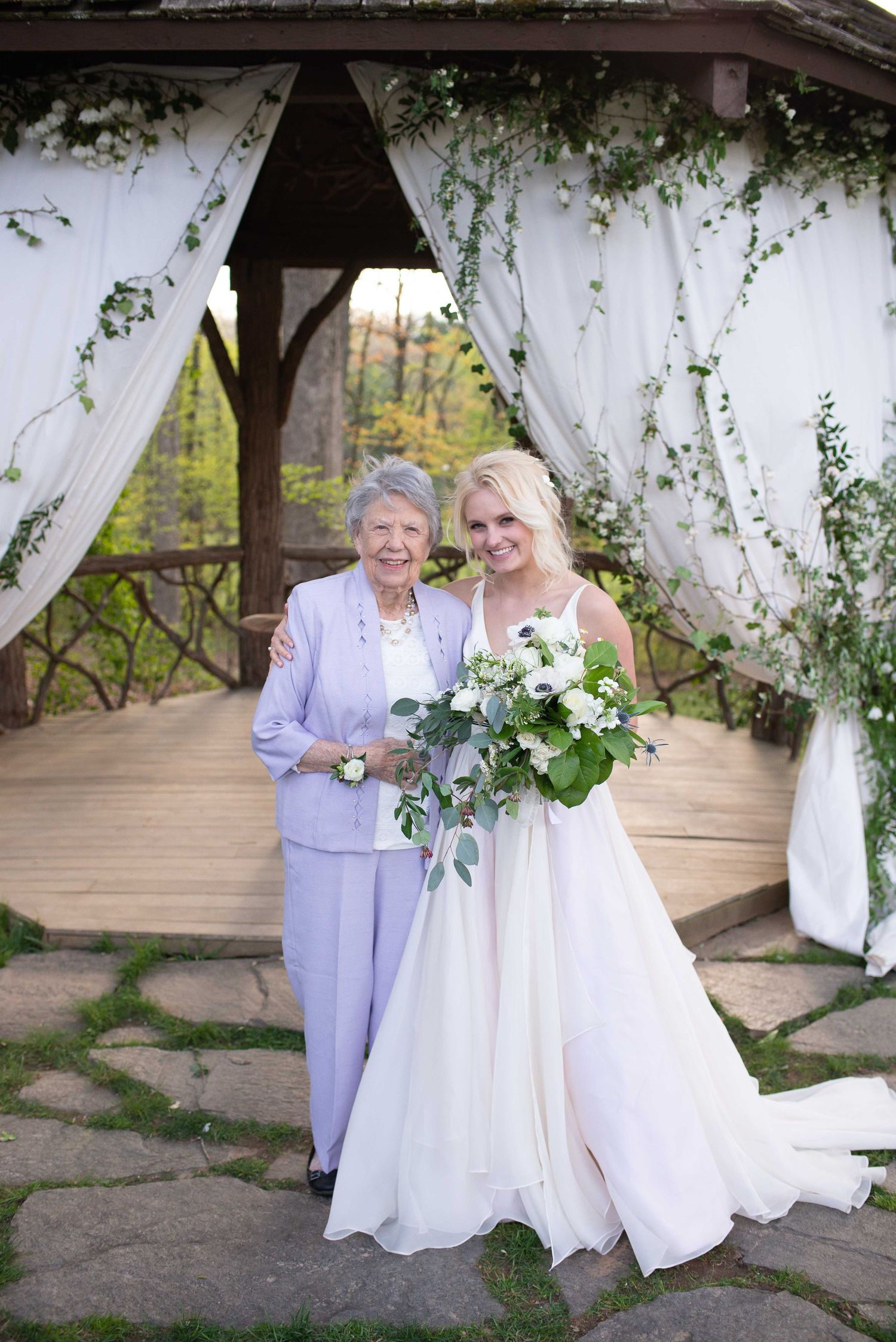 Bride in white gown holds bouquet beside elderly woman in lavender suit under floral gazebo.