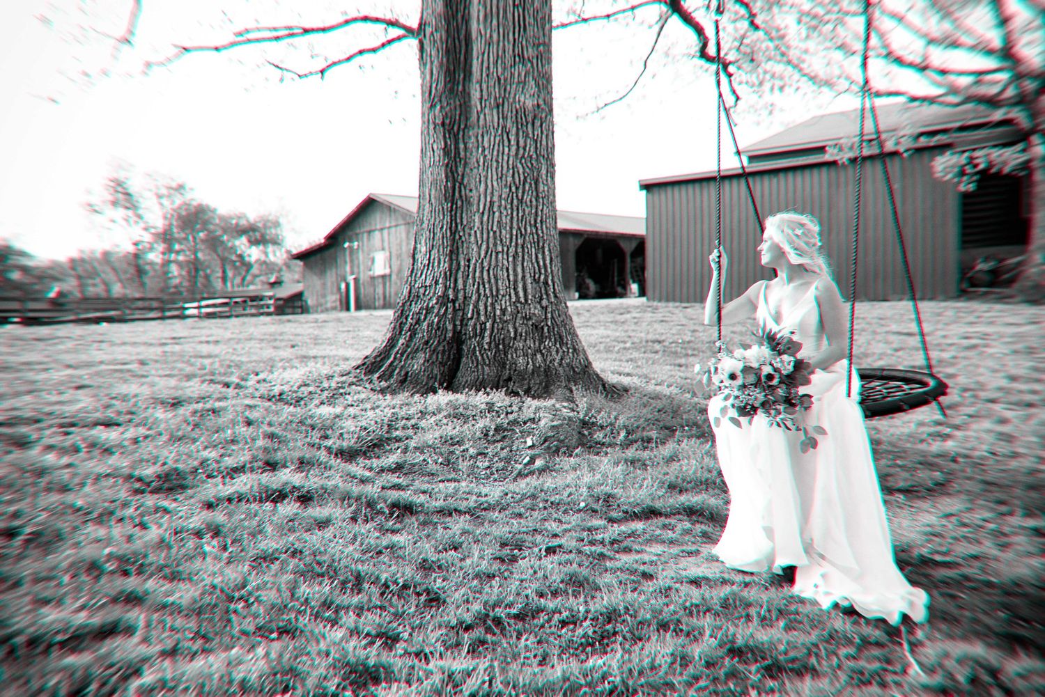 Bride in white gown standing by a large tree with a swing, holding a bouquet, with a barn in the background.
