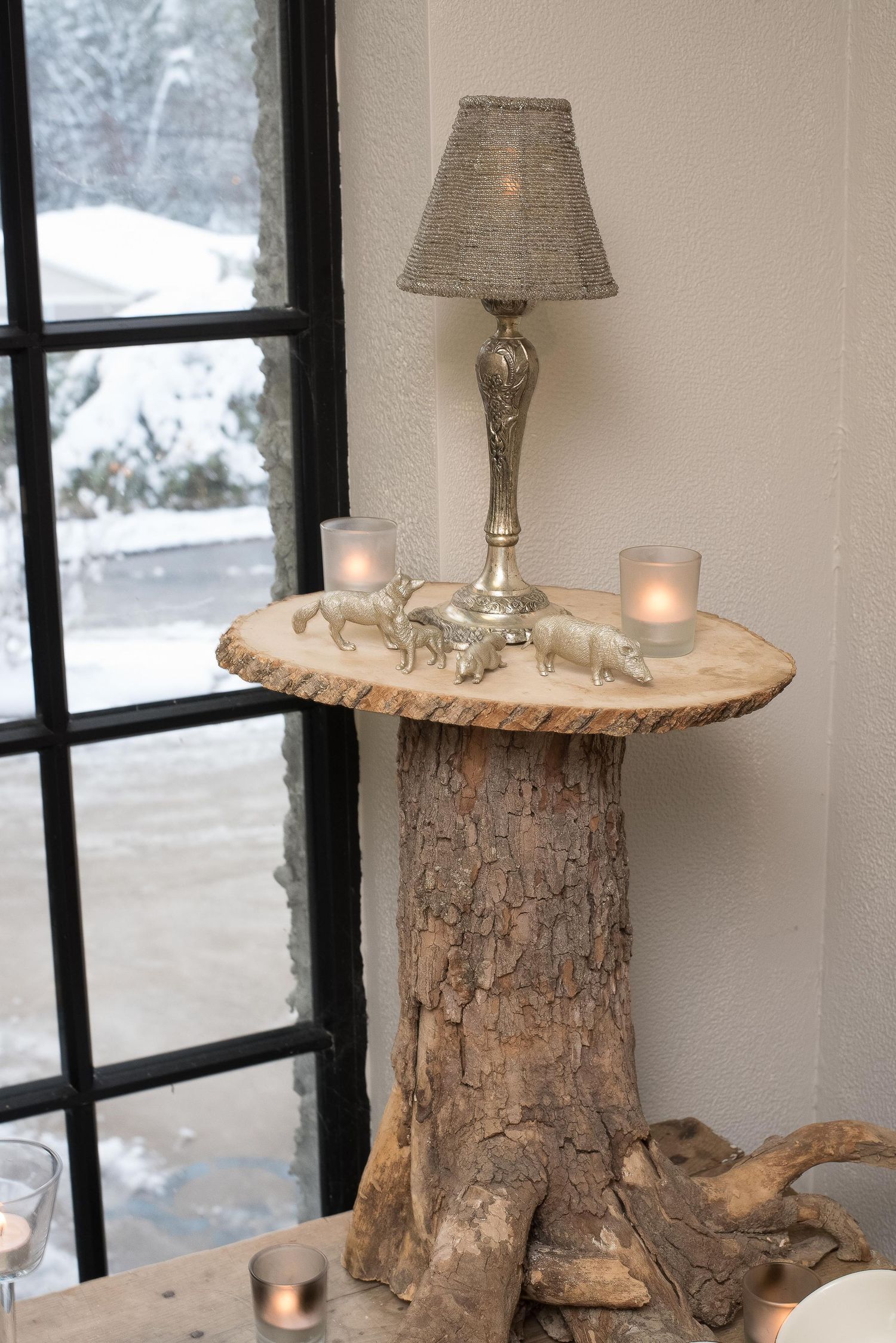 Rustic tree stump side table topped with a silver lamp, candles, and animal figurines near a snowy window.