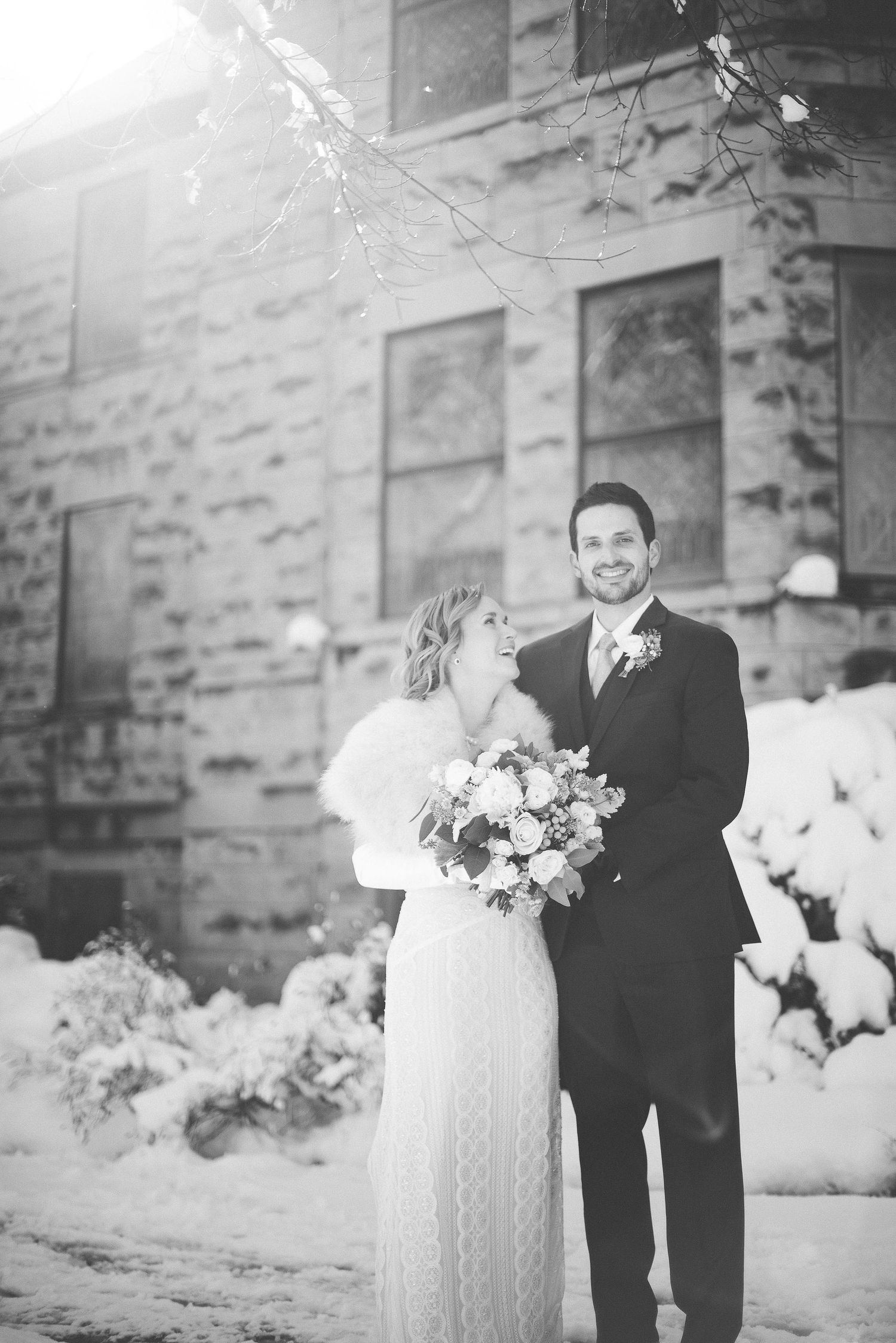 Black and white winter wedding photo of smiling bride and groom standing in snow outside stone building.