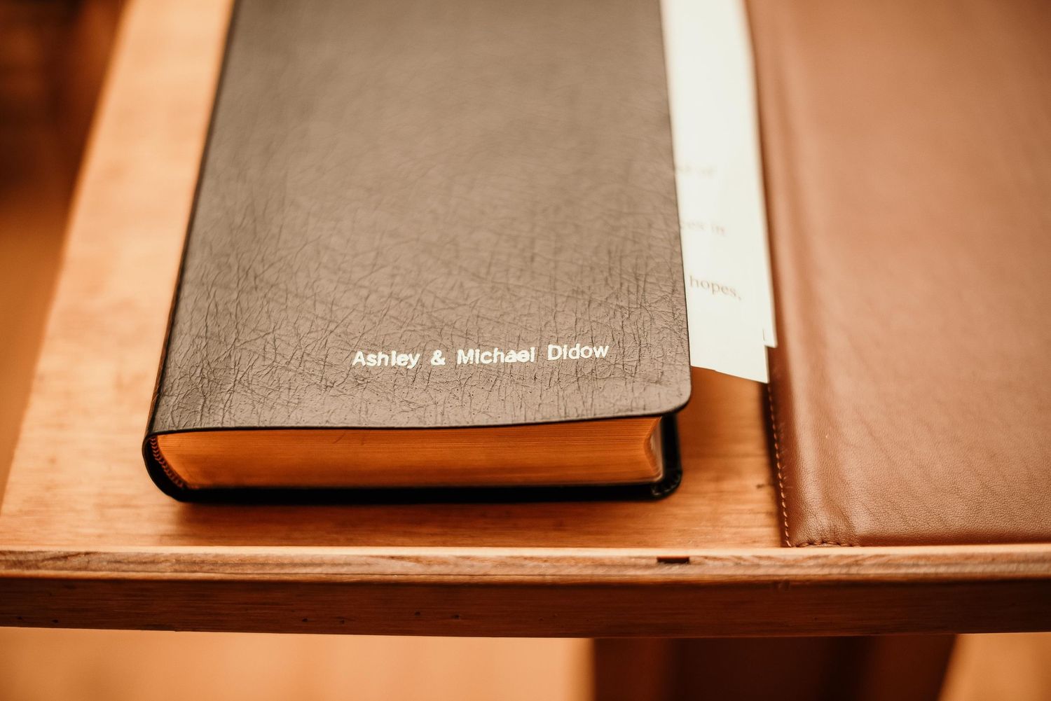 Personalized black leather wedding guest book embossed with Ashley & Michael Didow on a wooden table.