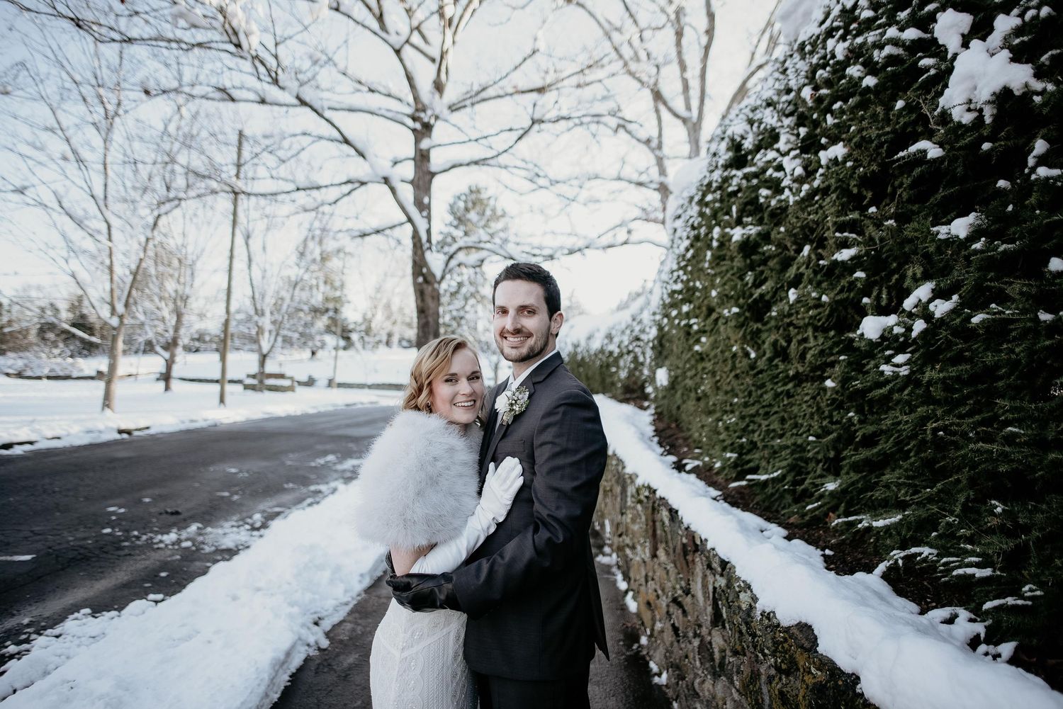 Bride and groom embracing outdoors in winter snow, bride wearing fur wrap and wedding dress beside stone wall.