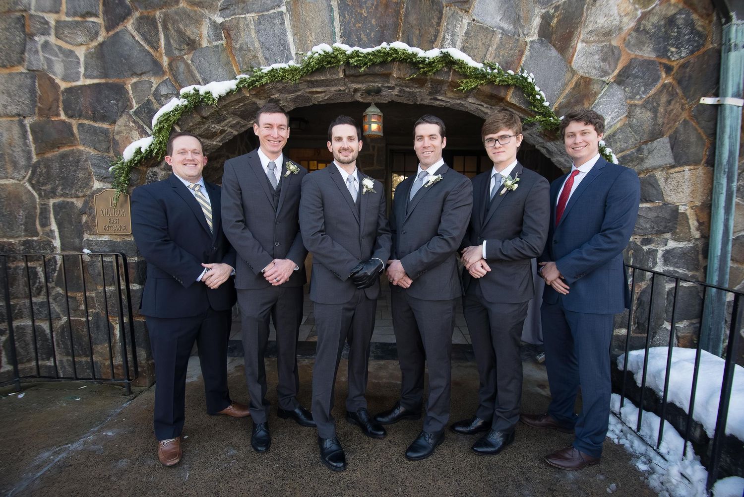Five groomsmen in gray suits pose together outside a stone church entrance decorated with greenery at a winter wedding.