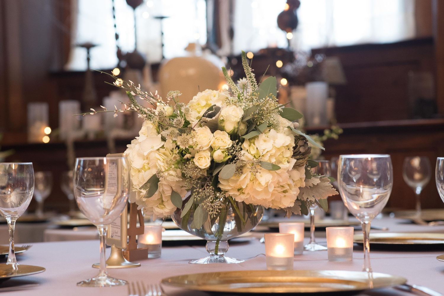 Elegant wedding centerpiece with white hydrangeas and greenery in a glass vase, surrounded by candles and gold charger plates.