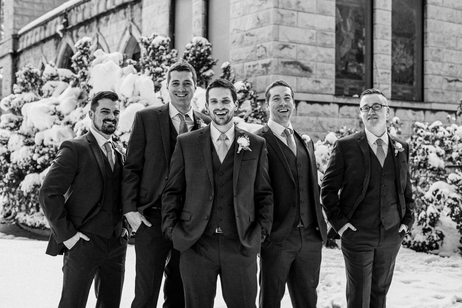 Black and white photo of a groom and four groomsmen in suits posing outdoors in snow at a winter wedding.