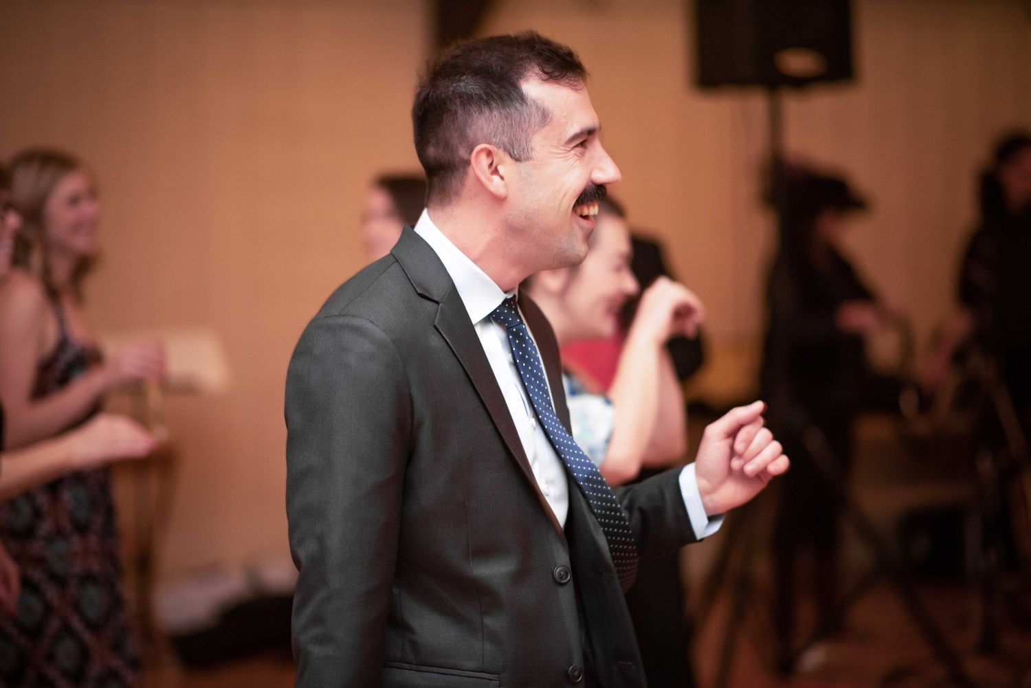 Man in gray suit and tie laughing at an indoor event or wedding reception.