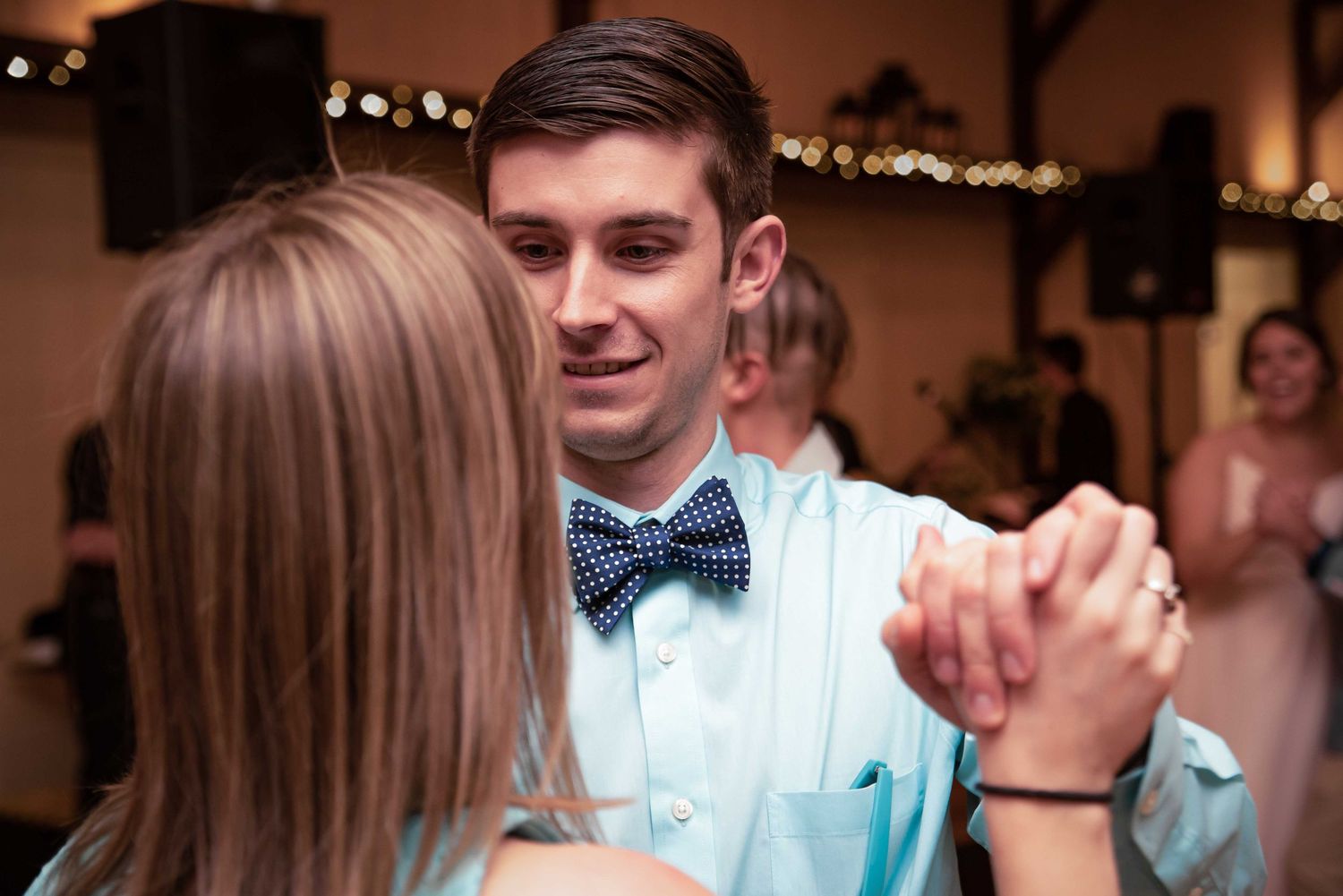 Young man in light blue shirt and navy bow tie shares a slow dance at a wedding reception.
