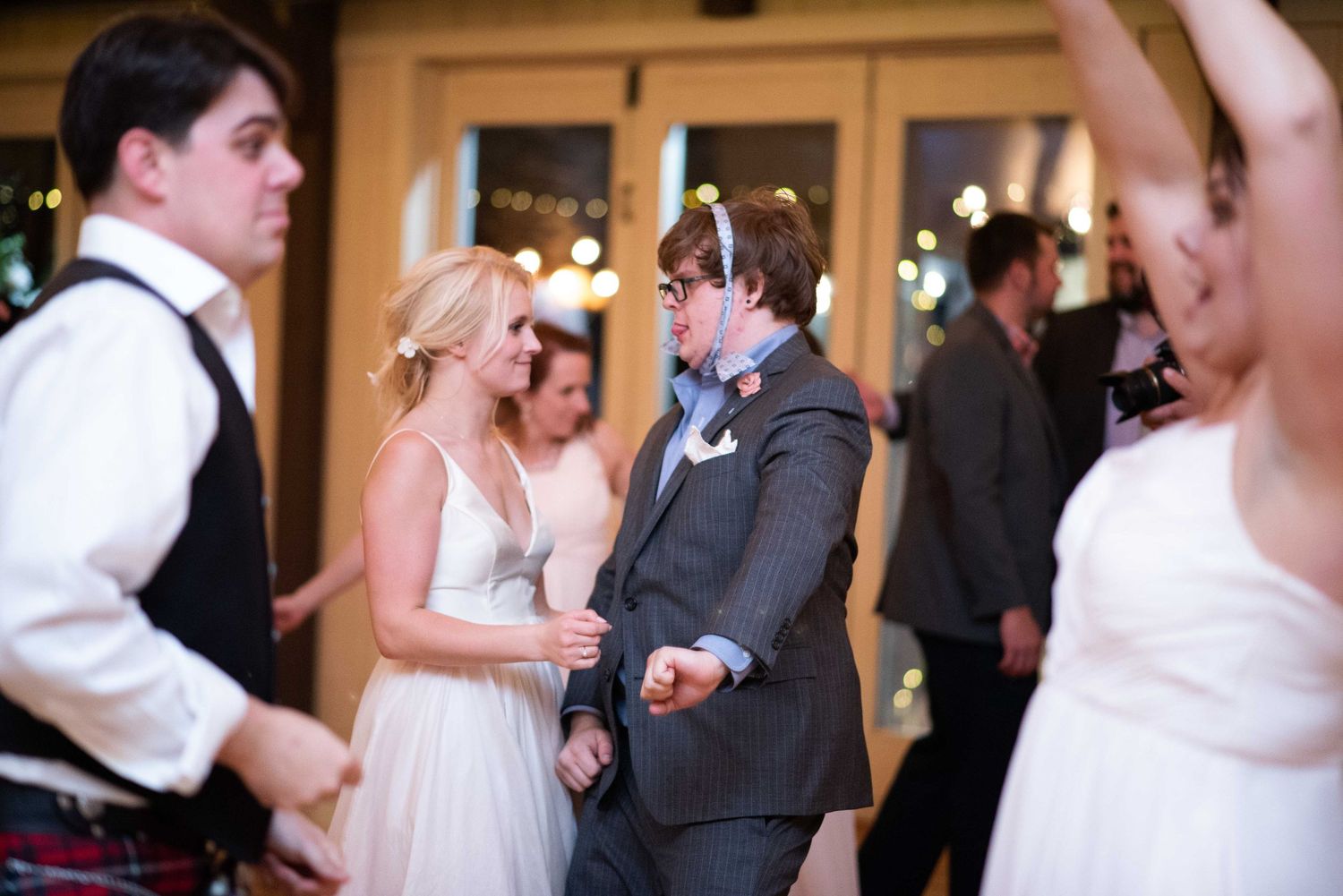 Guests dancing joyfully at a wedding reception, with bride in white gown and groom in gray suit on the dance floor.