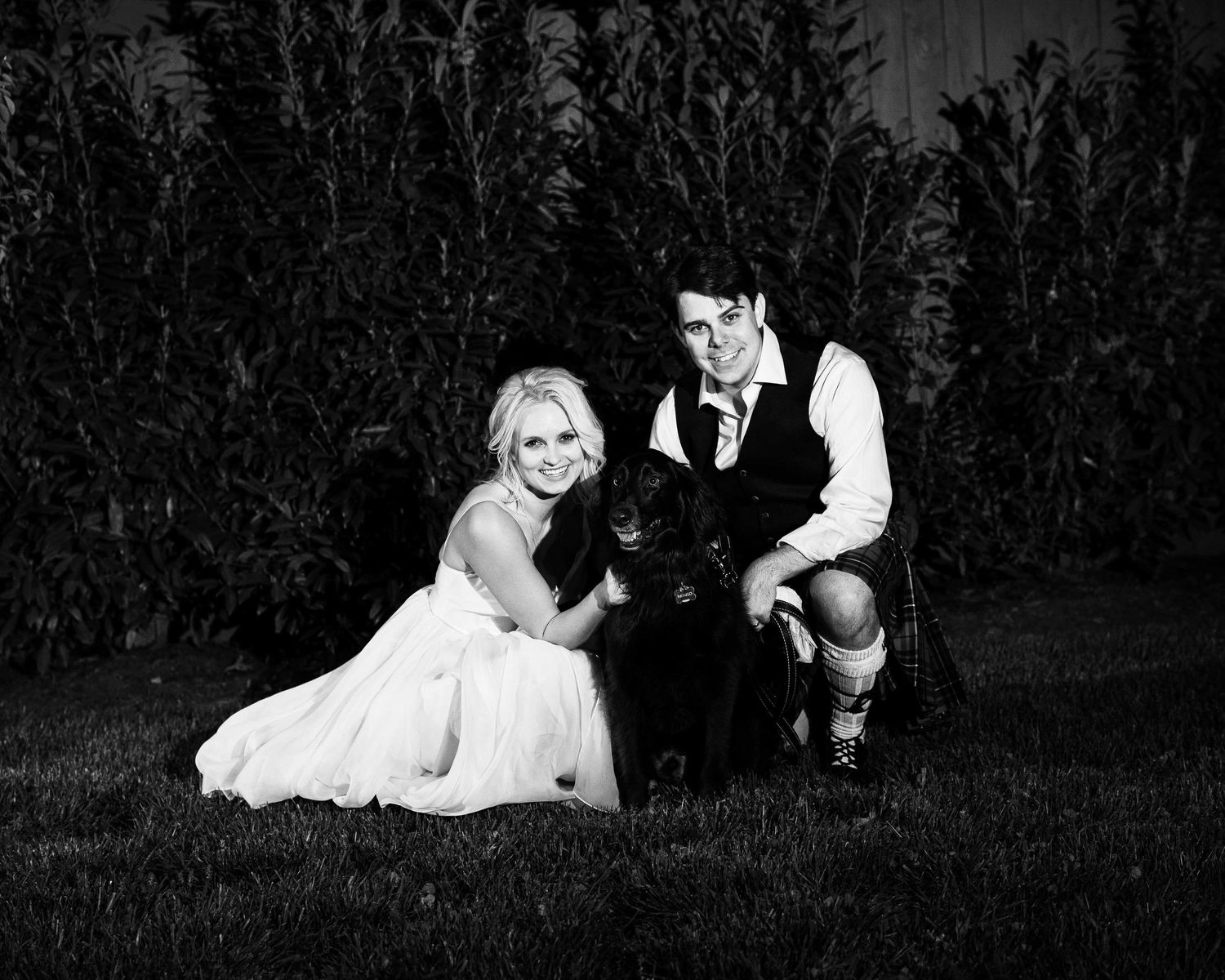 Black and white wedding photo of bride and groom posing with black dog on grass near cornfield.
