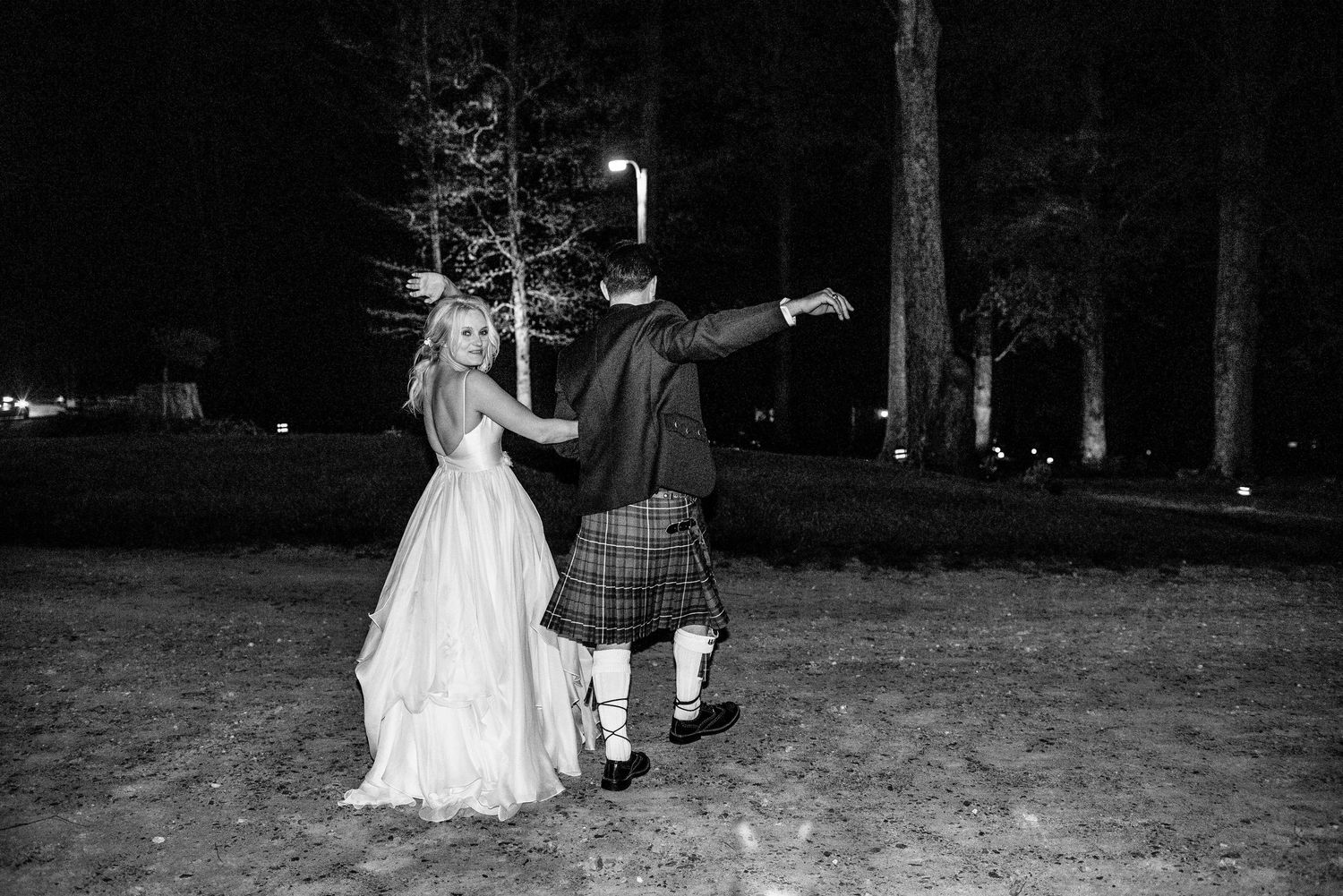 Black and white photo of a bride in a white gown and groom in a kilt dancing outdoors at night.