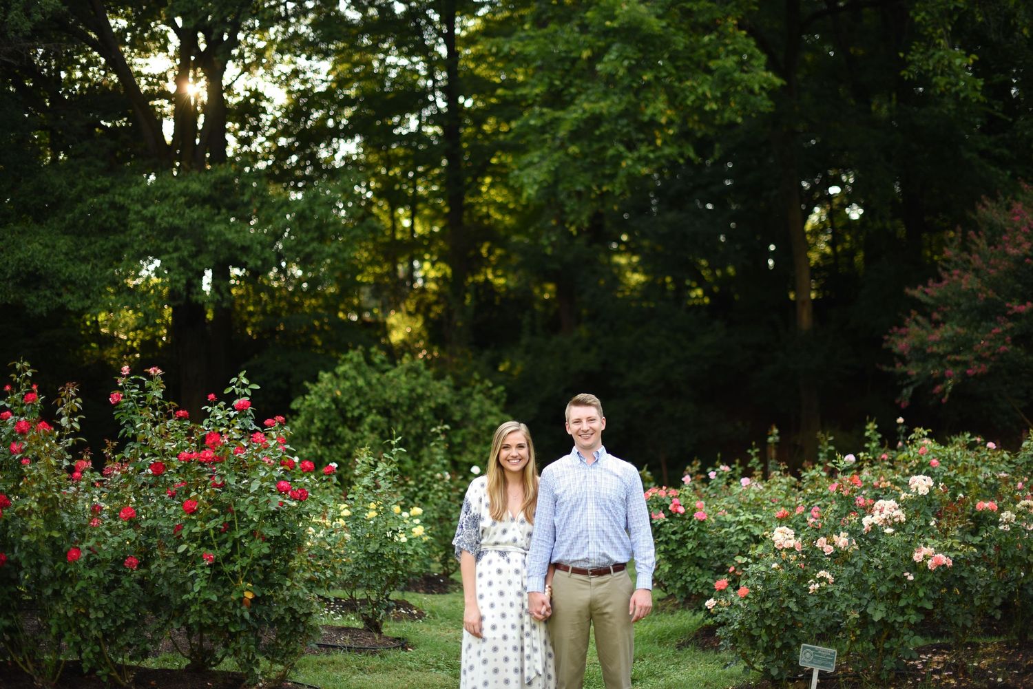 Smiling couple standing together in a vibrant rose garden surrounded by colorful blooms and tall trees.