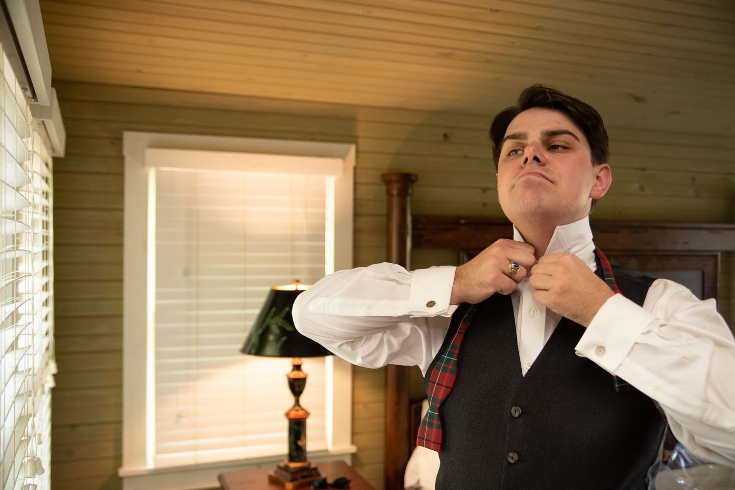 A groomsman adjusting his tie wearing a tartan vest and white dress shirt in a rustic room.
