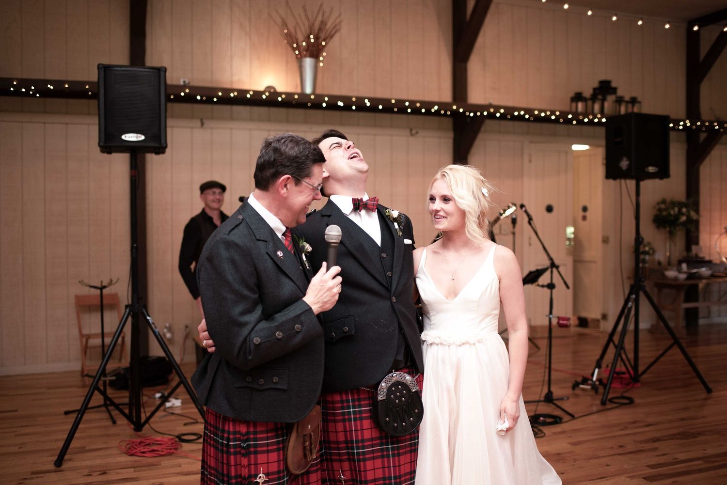 Groom in Scottish tartan kilt laughing with best man during wedding reception toast while bride smiles nearby.