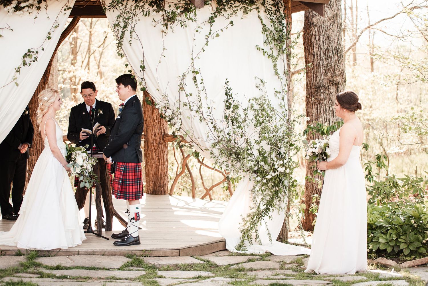 Outdoor wedding ceremony with bride and groom at rustic altar adorned with white draping and greenery, officiant in red tartan kilt.