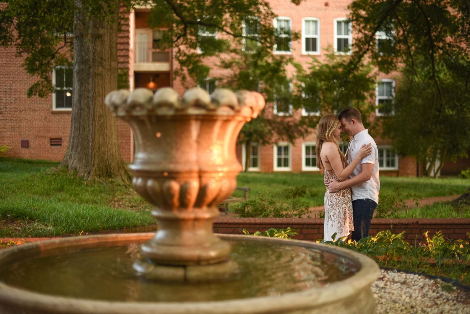 Couple embracing near an ornate stone fountain in a lush garden with a brick building backdrop at golden hour.