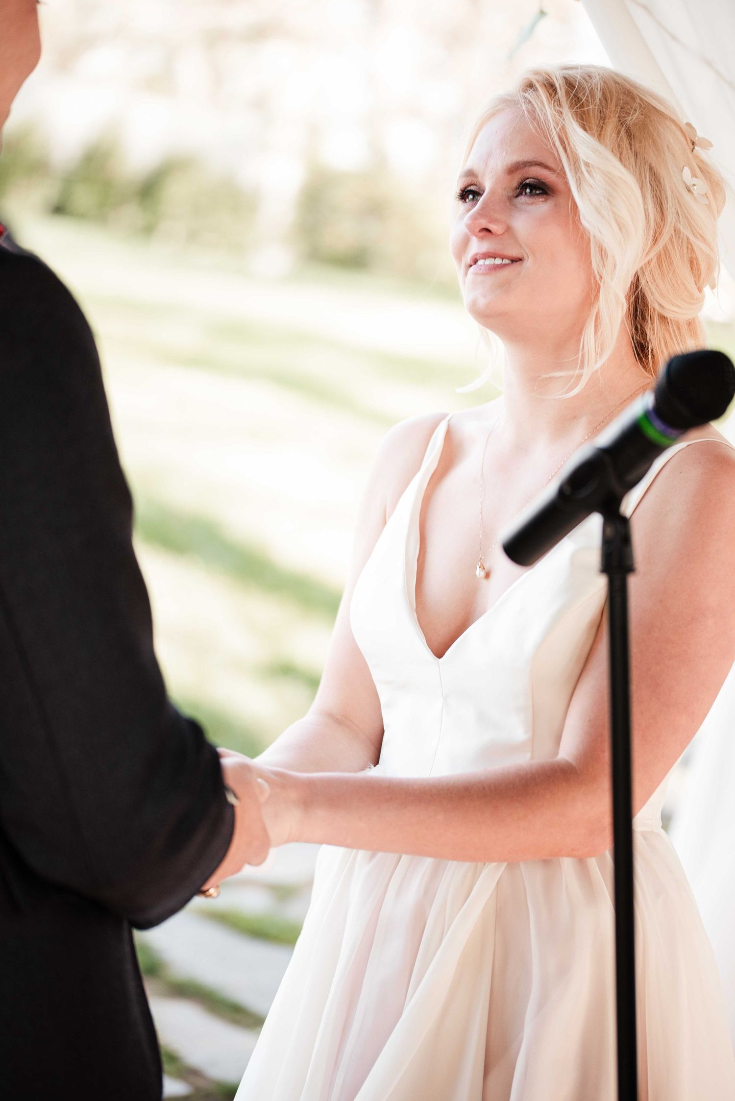 Smiling blonde bride in white gown holds hands with groom during outdoor wedding ceremony near microphone.