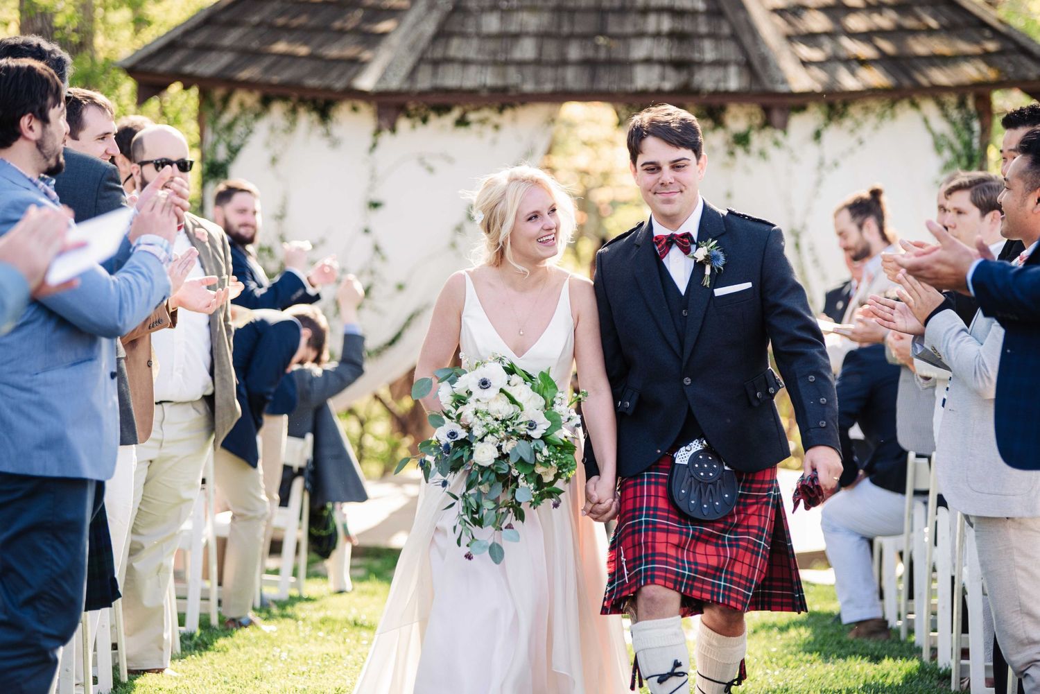 Bride and groom walk down the aisle, groom wearing a red tartan kilt, as guests applaud at an outdoor wedding ceremony.