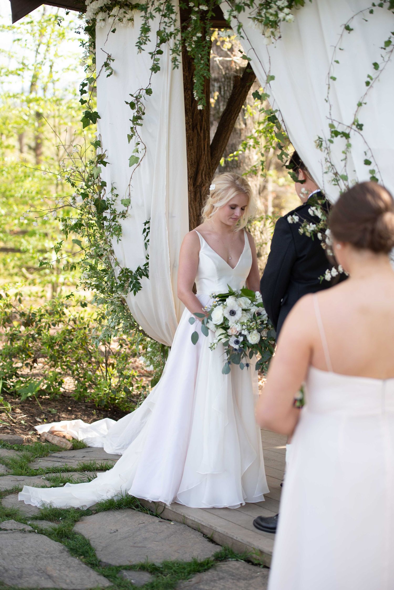 Bride in white V-neck gown holding bouquet stands under floral arch at outdoor wedding ceremony.