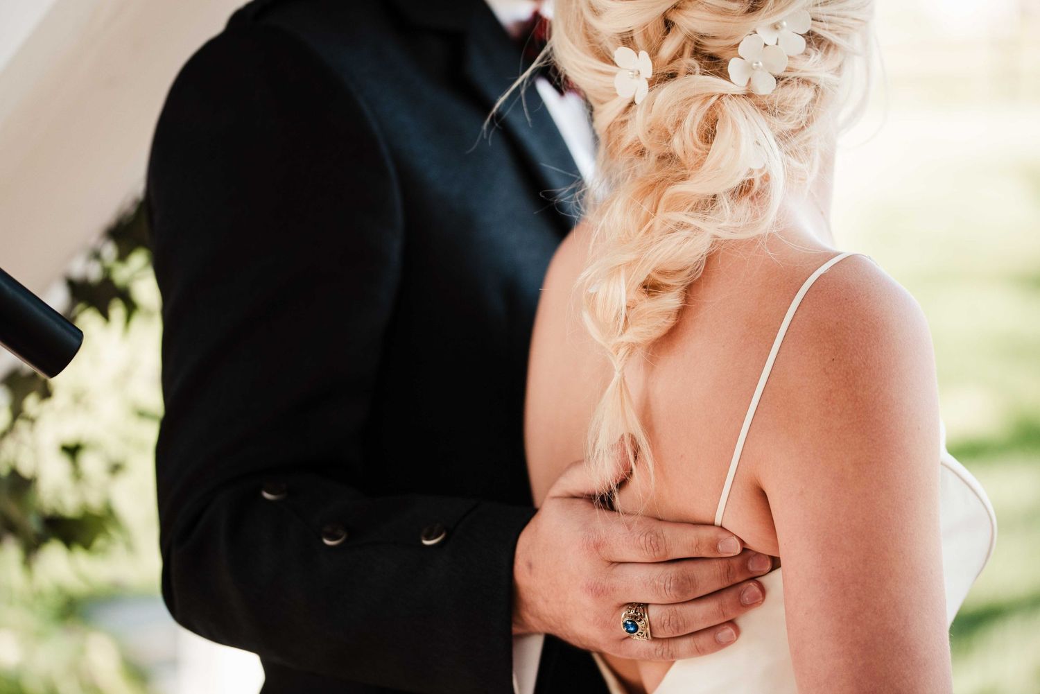 Groom in black tuxedo embracing bride in white wedding dress with elegant braided updo hairstyle outdoors.