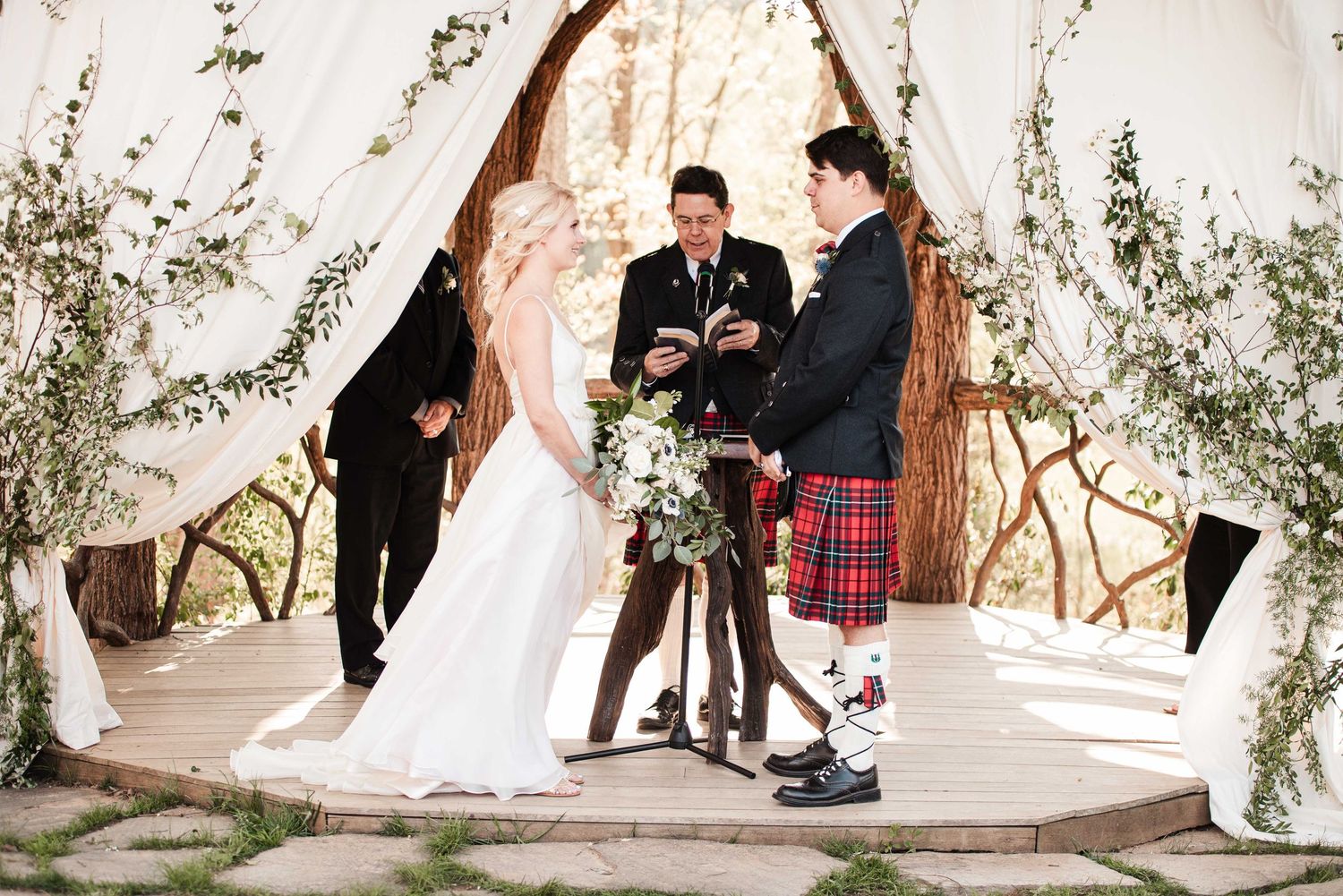 Outdoor wedding ceremony under a rustic wooden arch with white draping, bride in white gown, groom in red tartan kilt.