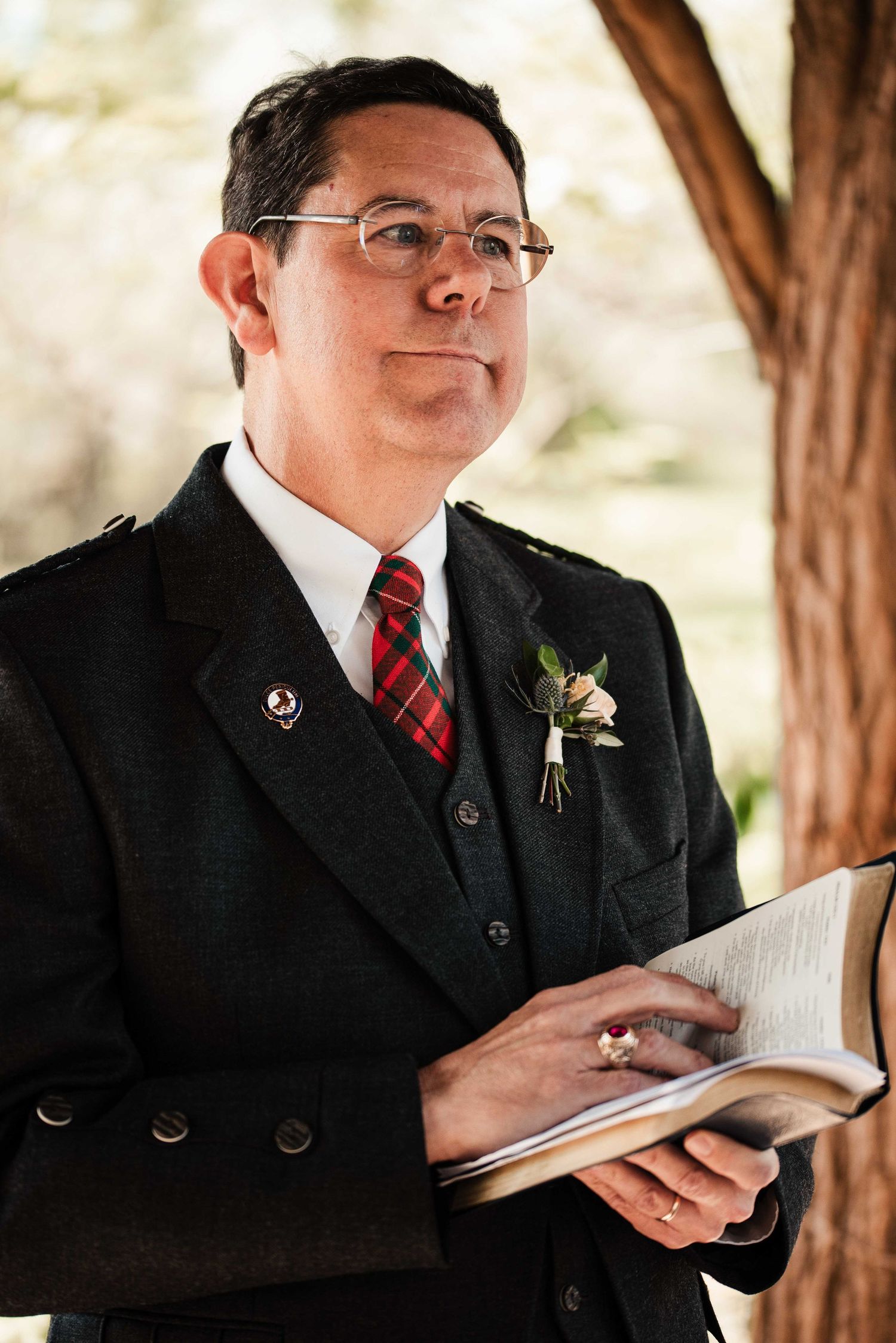 Man in dark suit with plaid tie and boutonniere holds open book outdoors, appearing to officiate a ceremony.