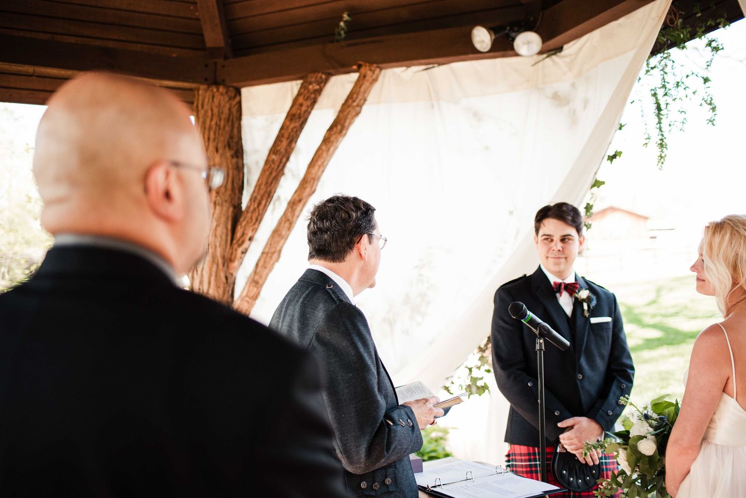 Groom in kilt stands at wedding altar under rustic wooden pavilion with white draping during outdoor ceremony.