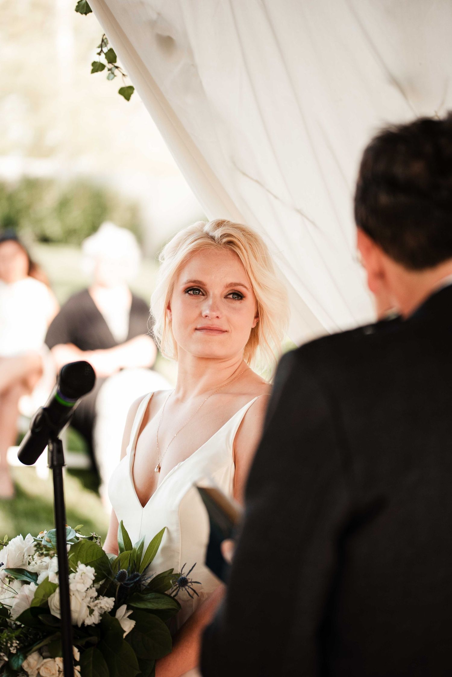 Bride in white V-neck gown gazes lovingly at groom during outdoor wedding ceremony under draped tent.