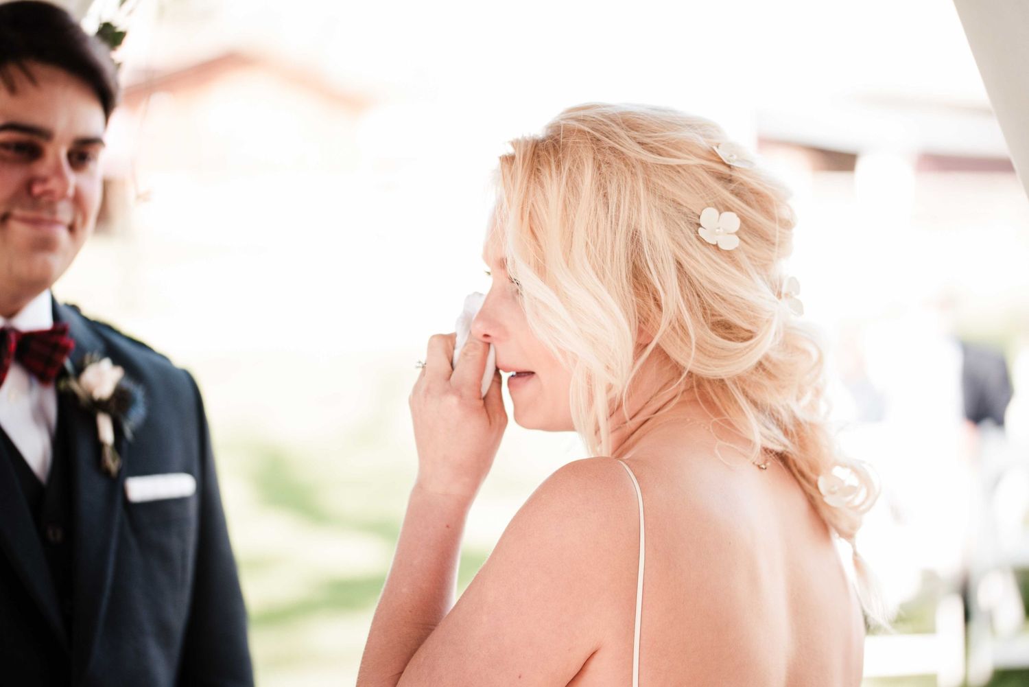 Tearful blonde bride wiping her eye while smiling during an emotional outdoor wedding ceremony.