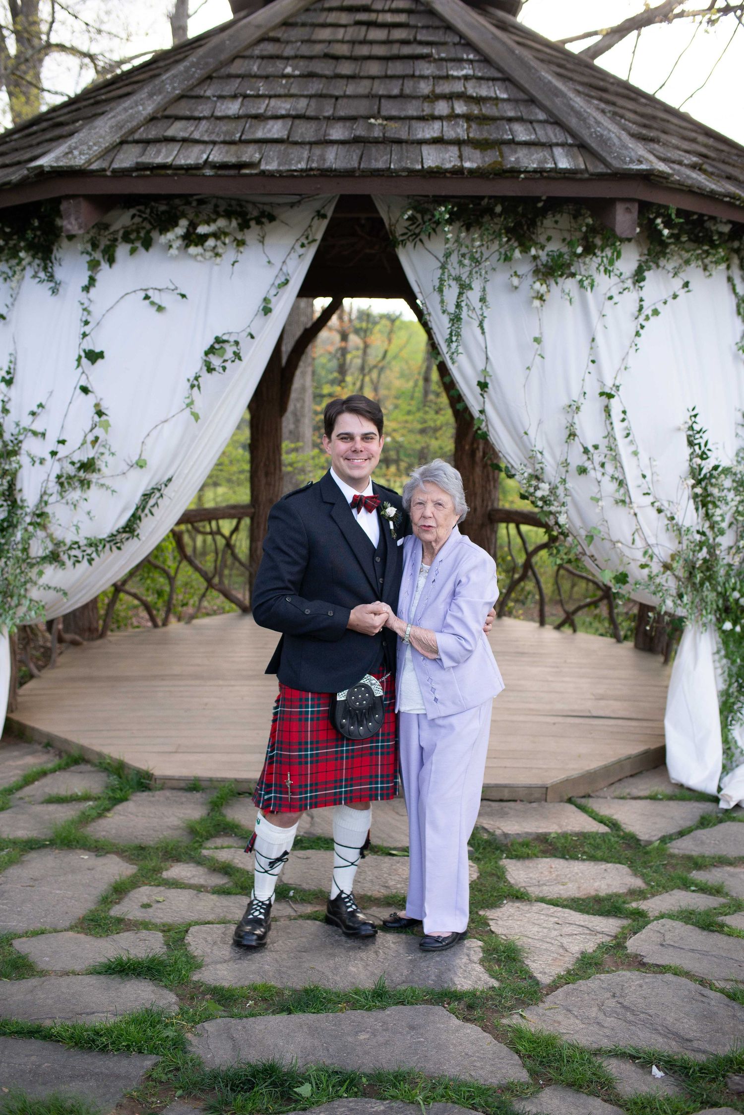 Groom in red tartan kilt and elderly woman pose together under decorated gazebo at outdoor wedding ceremony.