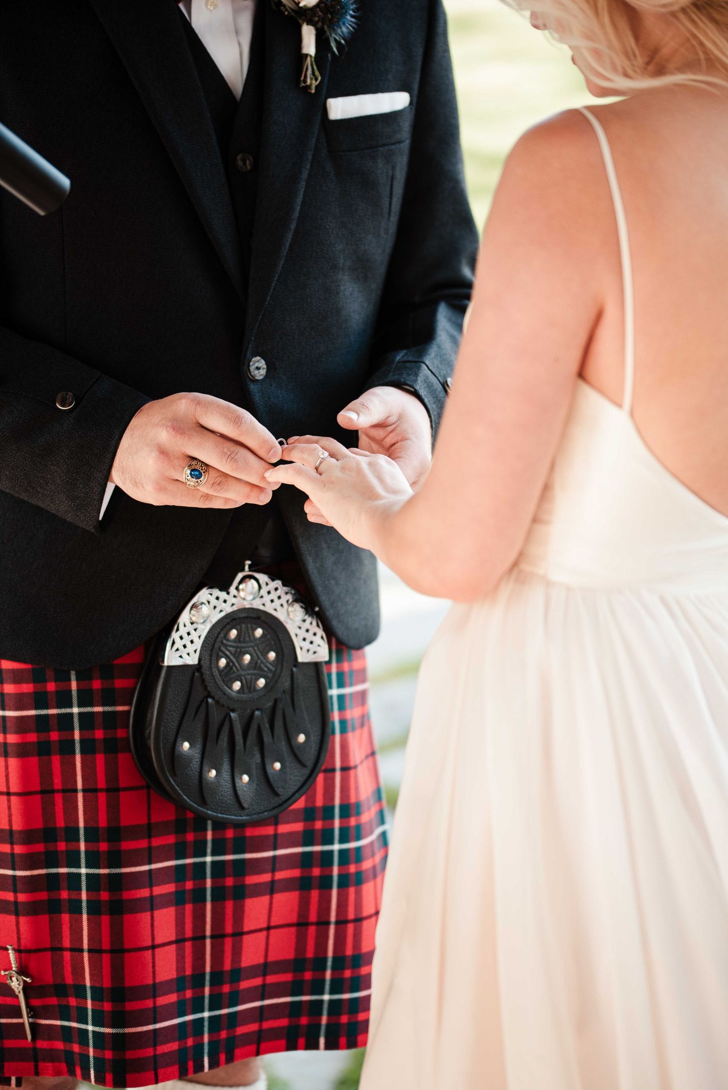 Groom in red tartan kilt and sporran exchanges wedding rings with bride in white dress during ceremony.