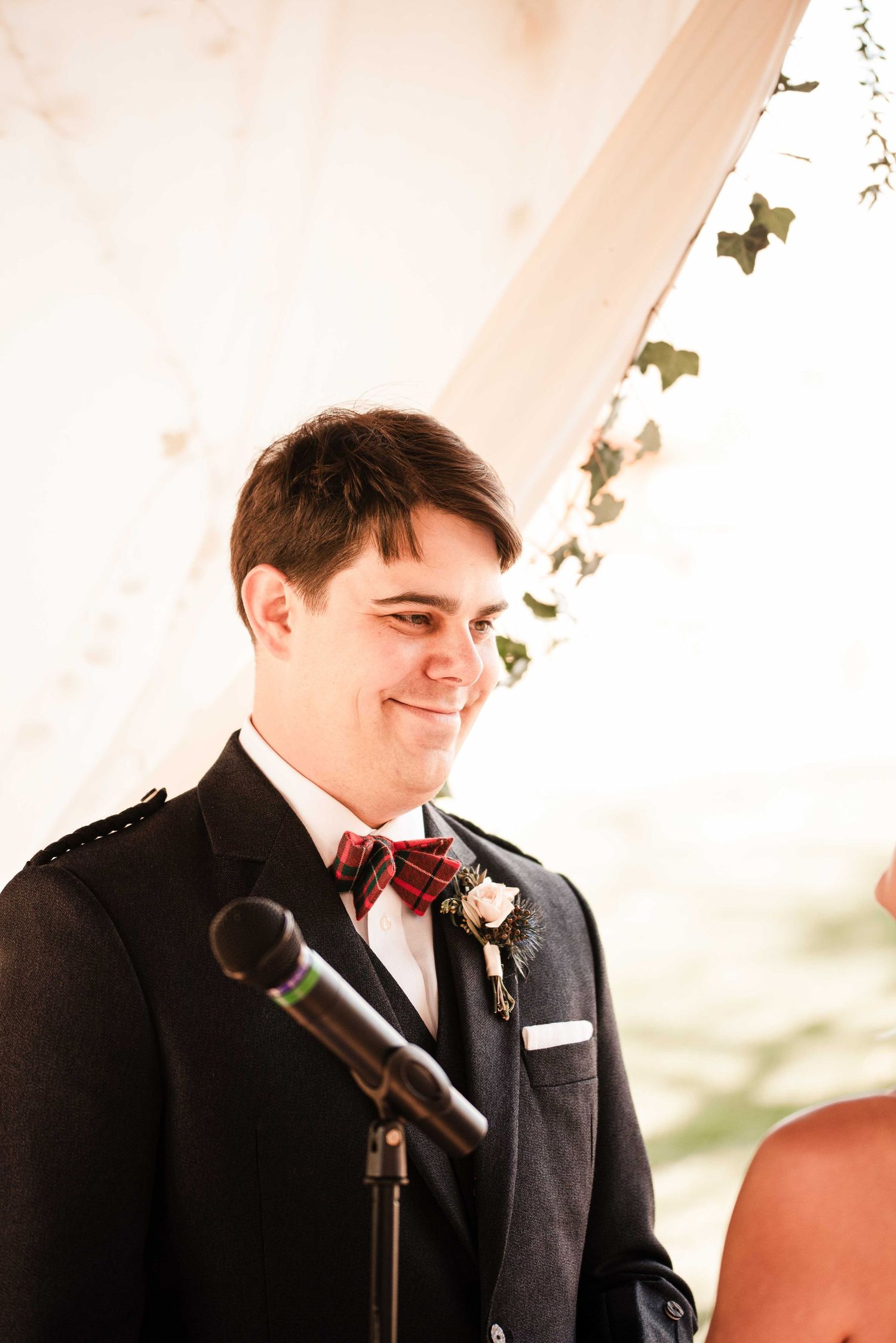 Smiling groom in dark suit with plaid bow tie and boutonniere at wedding ceremony altar.