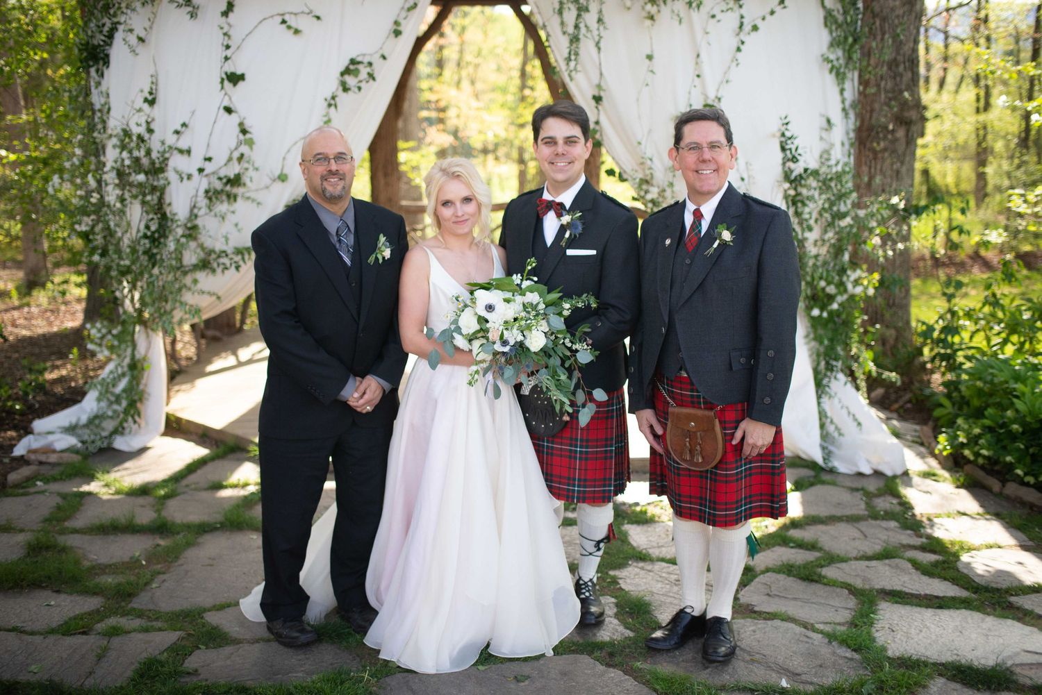 Wedding party poses outdoors with bride in white gown, groom in tuxedo, and man in traditional red tartan kilt.