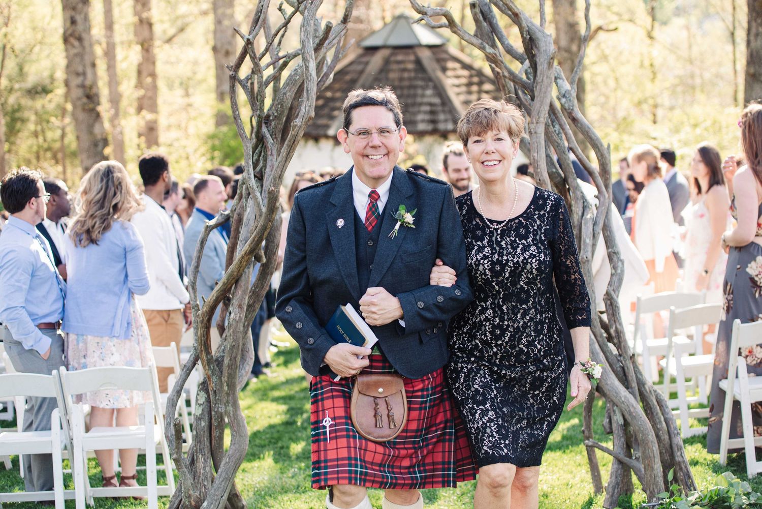 Man in Scottish kilt and woman in floral dress smile under rustic arch at outdoor wedding ceremony.