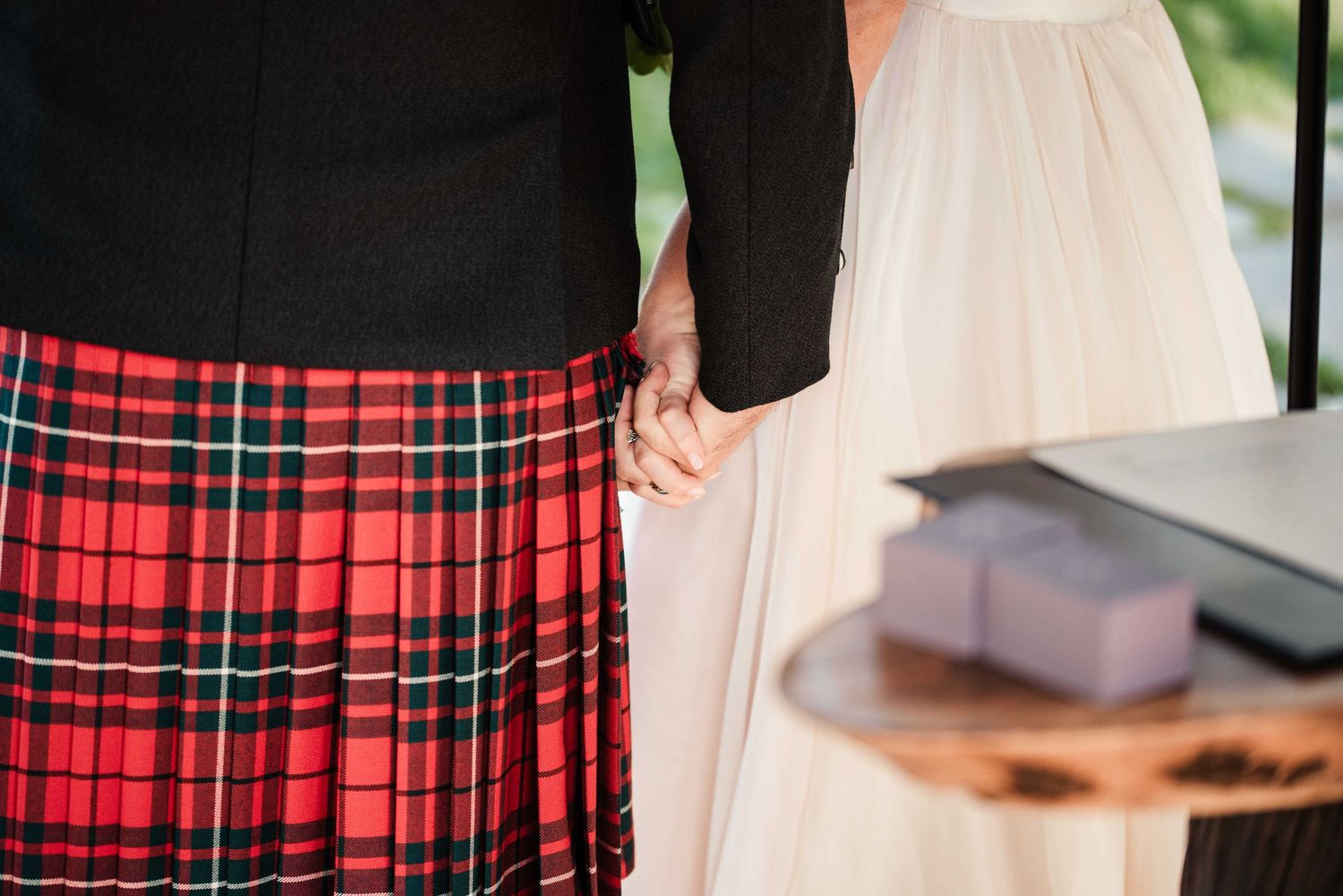 Couple holding hands at wedding ceremony, groom wearing traditional red tartan Scottish kilt, bride in white dress.