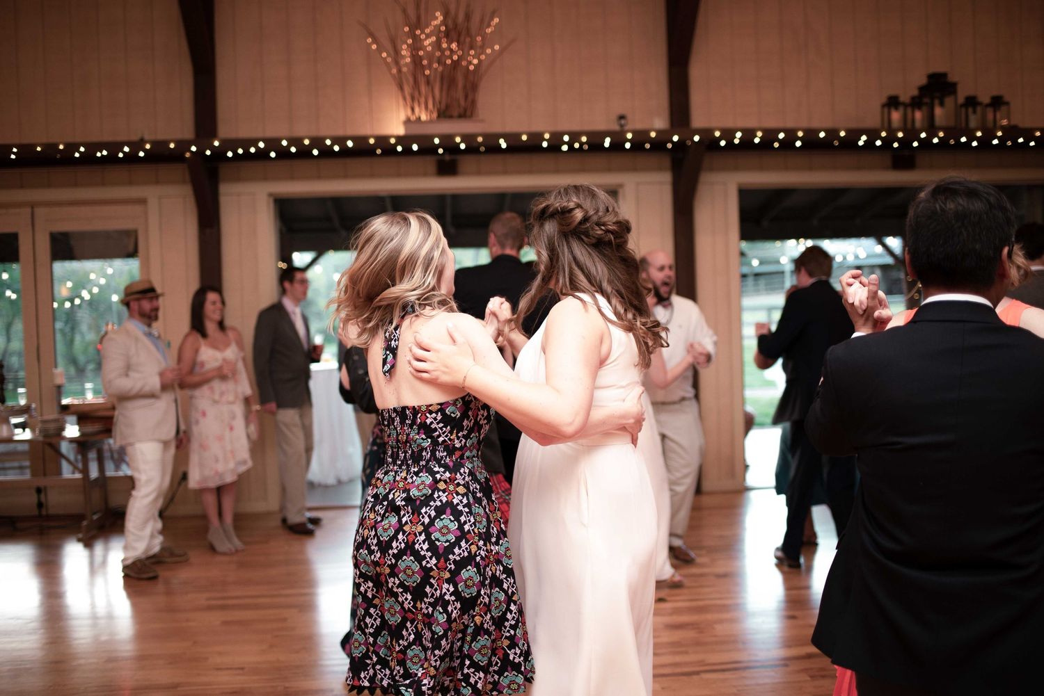 Two women embrace on a wooden dance floor at an elegant indoor wedding reception with string lights.