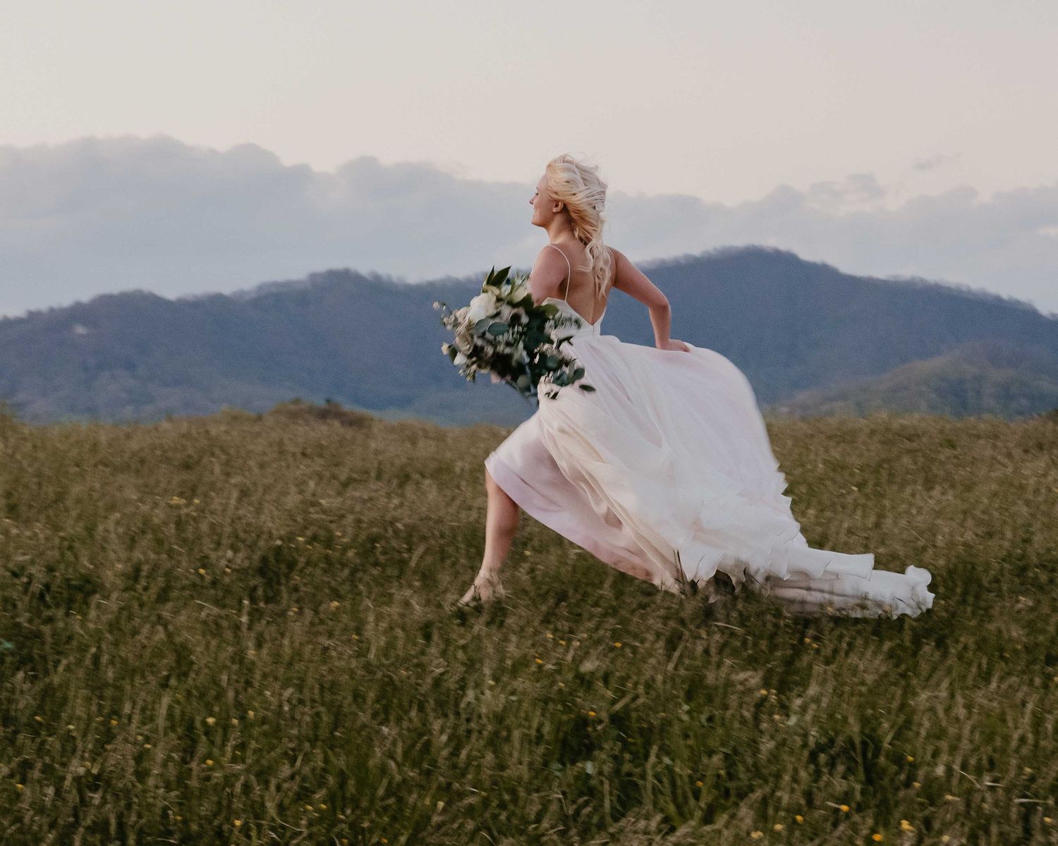 Bride in white gown running through mountain meadow holding bouquet, with rolling hills and clouds in background.
