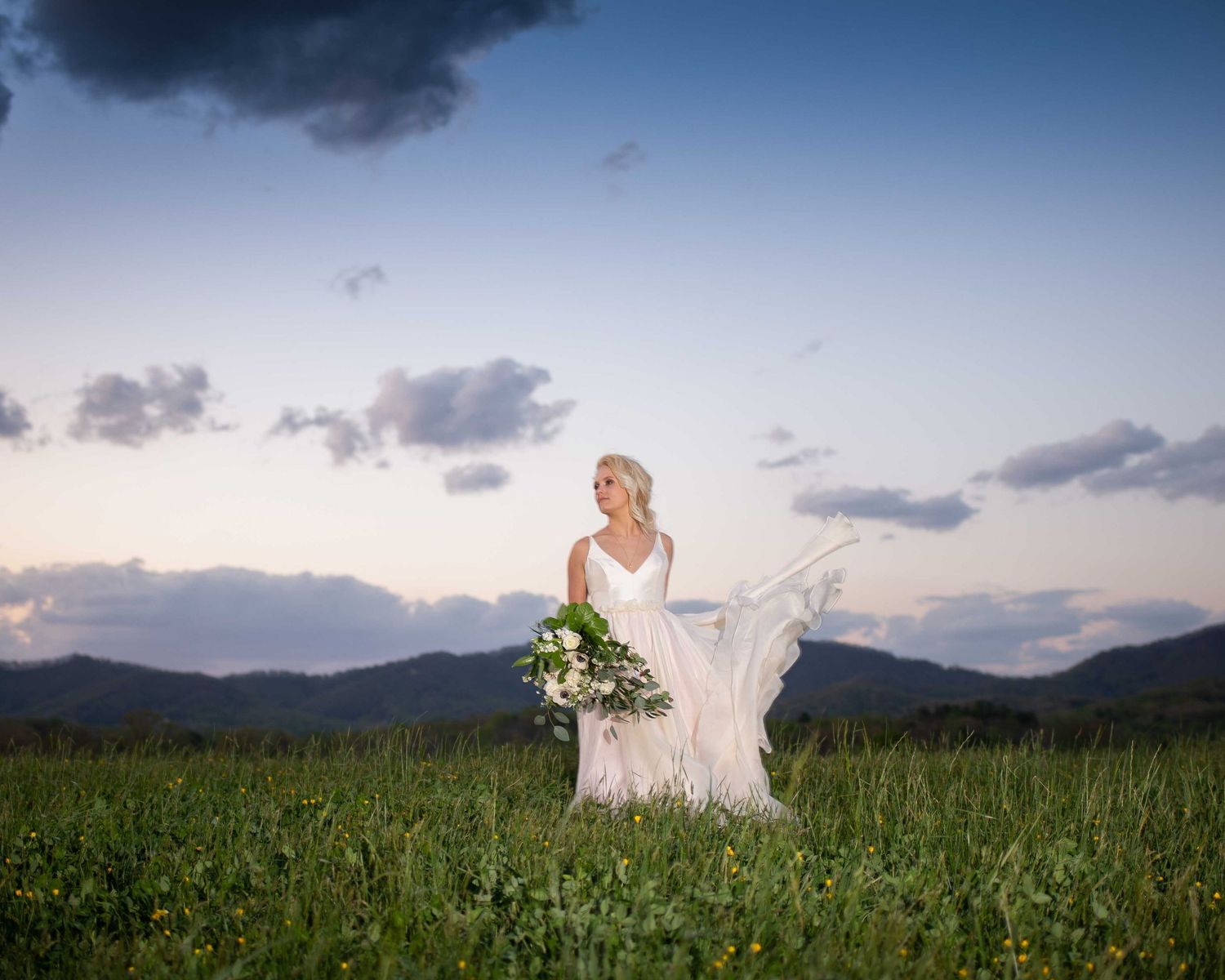 Bride in white gown holding bouquet stands in green meadow with mountain backdrop at dusk.
