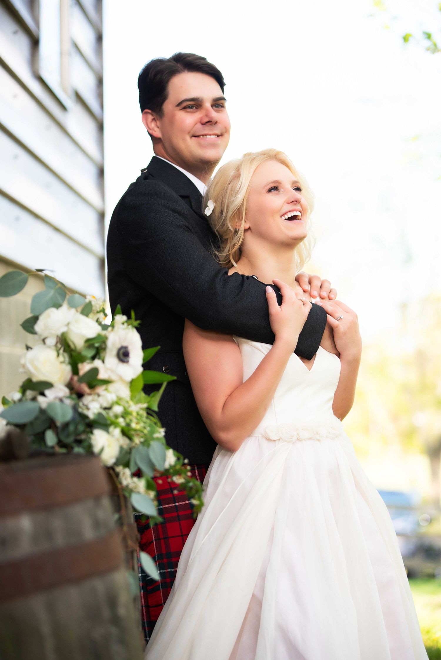 Smiling bride in white gown and groom in black jacket with red tartan kilt embrace outdoors near white building.